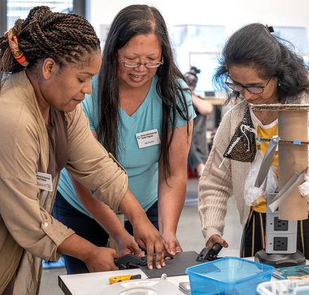 Three educators collaborate in a bright workshop, using clamps and cardboard to build a structural model during a teacher program session