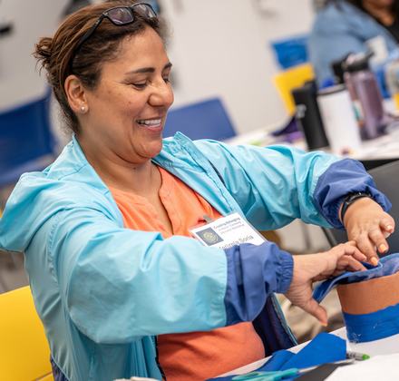A smiling educator wearing a blue jacket participates in a hands-on maker-centered workshop, assembling a project with blue fabric and tape