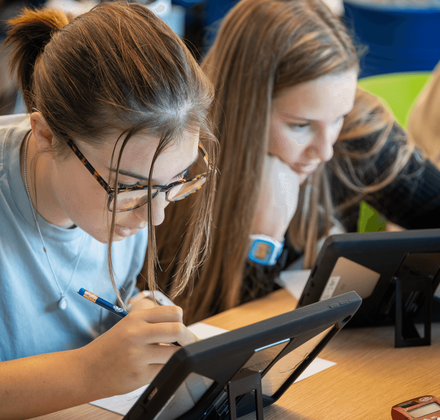 Two students leaning over tablets and writing on paper at a classroom table, focused on their work