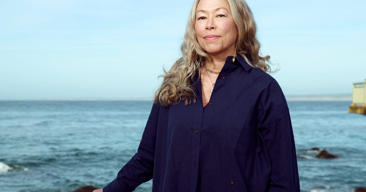 A woman with blonde brown hair wearing a blue button up top stands in front of an ocean background