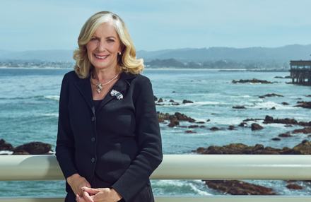A woman with blonde hair in a black blazer smiles while standing in front of an ocean background
