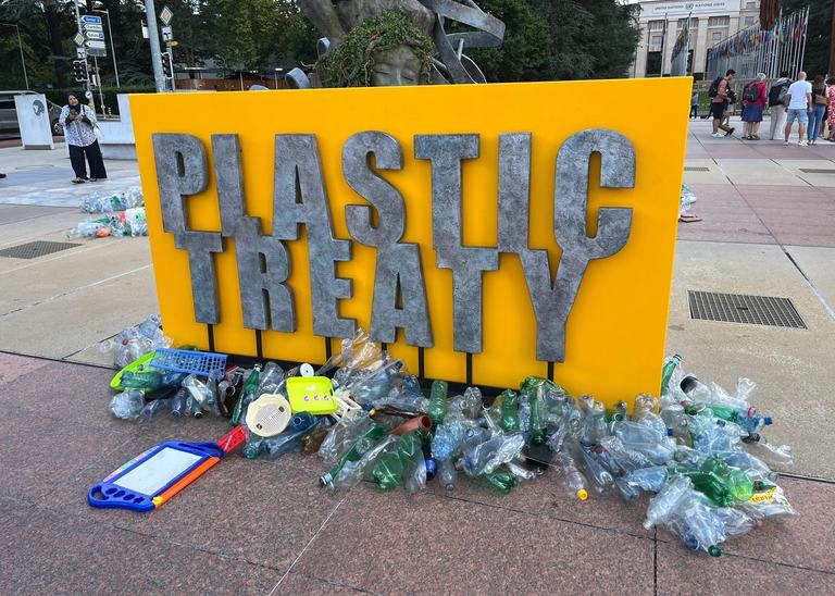 Large yellow sign with plastic treaty written on it in front of plastic trash.