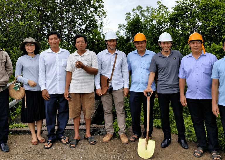 A group of nine adults standing outdoors in a lush green area, some wearing construction helmets and holding a shovel, posing together on a dirt path