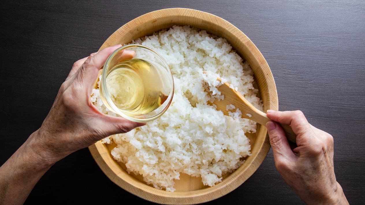 Hands mix vinegar into cooked sushi rice in a round wooden bowl
