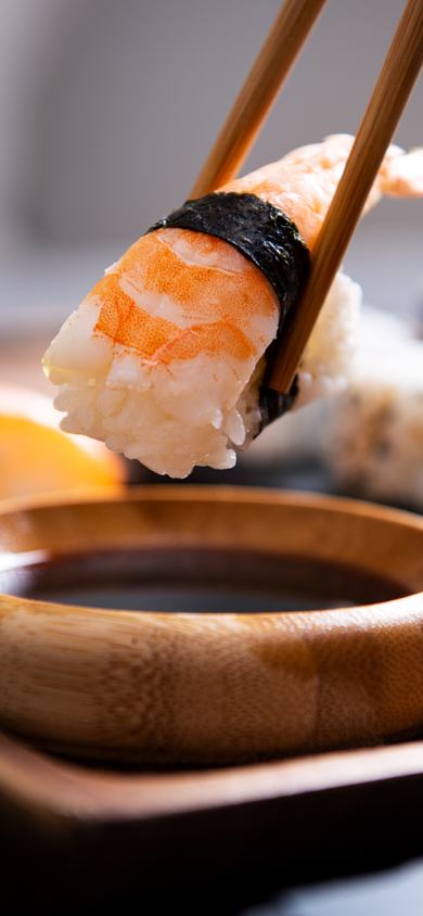 Chopsticks holding shrimp sushi over a small dipping sauce bowl with assorted sushi rolls in background