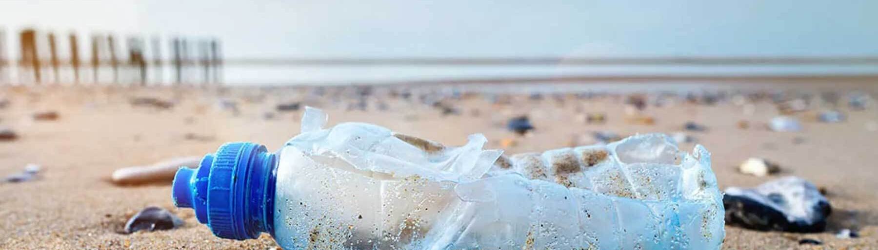 Crushed plastic water bottle with a blue cap littering a sandy beach, with ocean waves and wooden posts visible in the background