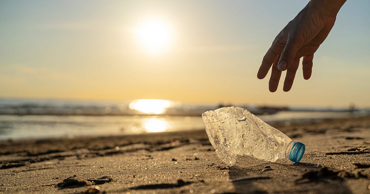 Hand reaching down to pick up a crushed plastic bottle on a sandy beach at golden sunset, with ocean and glowing sun in the background