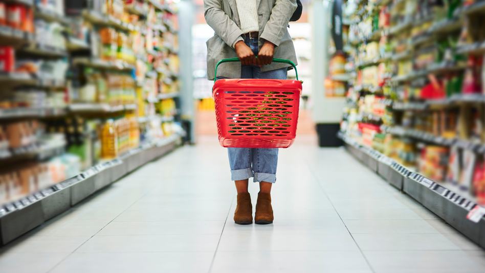 A person standing in a supermarket aisle holding a red shopping basket with both hands